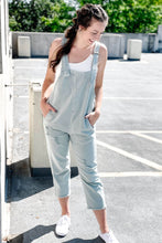 Load image into Gallery viewer, Women&#39;s distressed relaxed fit denim blue overalls paired with a cropped white ribbed tank top and universal threads white sneakers. 
